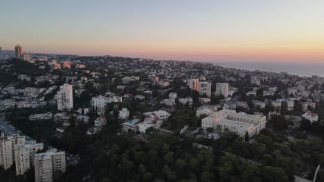 Rising-aerial-view-of-a-city-full-of-buildings-and-trees-at-sunrise,-Haifa,-Israel