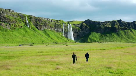 Fotógrafos-De-Naturaleza-Caminando-Sobre-Un-Campo-De-Hierba-Verde-Hacia-Seljalandsfoss-En-Islandia