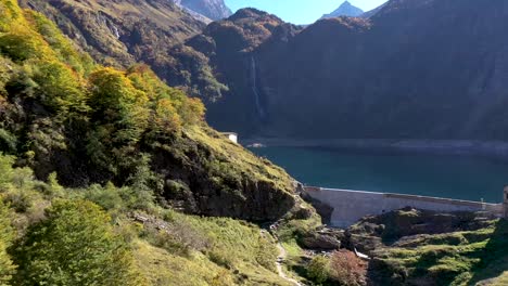Lac-d'Oô-artificial-lake-in-the-French-Pyrenees-with-hikers-standing-near-the-dam-wall-on-a-ridge,-Aerial-approach-shot