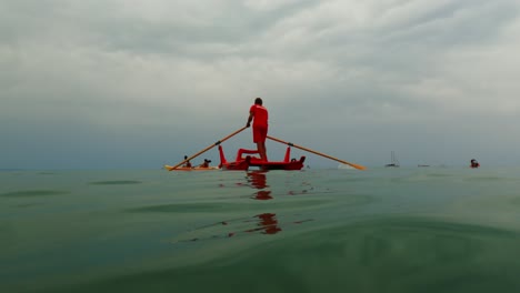 Red-Italian-lifeguard-ready-for-first-aid-emergency-standing-and-rowing-rescue-boat-while-watching-people-on-kayak-and-sup-in-background
