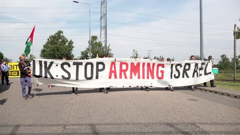 Protestors-march-holding-a-large-white-banner-that-says,-“UK-Stop-Arming-Israel”-at-a-protest-against-the-DSEi-arms-fair