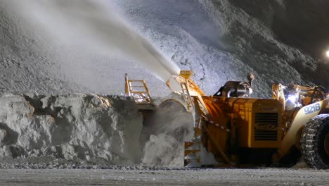 Lader-Montierte-Schneefräse-In-Betrieb,-Um-Schnee-Oben-Auf-Schneehaufen-Zu-Blasen,-Während-Ein-Traktor-Nachts-Vorbeifährt