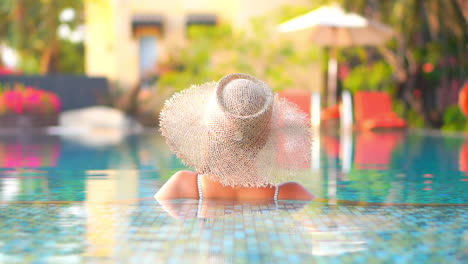 Back-to-the-camera,-low-angle,-a-young-woman-wearing-a-straw-sun-hat-leans-up-along-the-edge-of-a-resort-swimming-pool