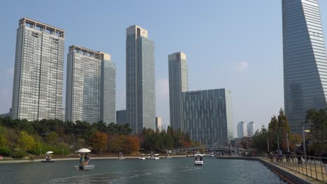 Posco-Tower-Und-Songdo-Central-Park-In-Incheon-Mit-Menschen,-Die-Mit-Booten-Reisen-Und-Während-Der-Covid-19-Mit-Gesichtsmasken-Am-See-Spazieren-Gehen,-Mit-Hochhäusern-Gegen-Klaren-Himmel