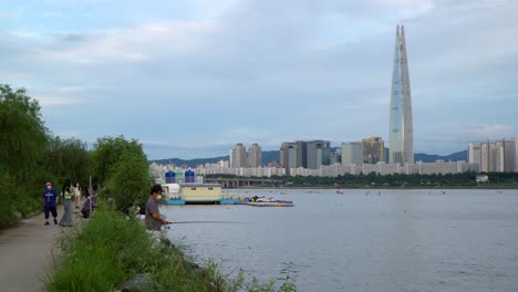 Man-Fishing-On-Han-Rivershore-With-Seoul-Cityscape-And-Lotte-World-Tower-In-South-Korea