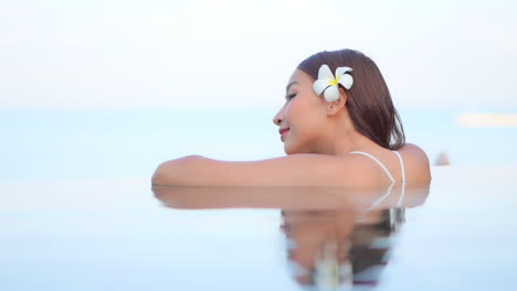 A-close-up-of-an-attractive-woman-with-a-flower-behind-her-left-ear-leans-along-the-edge-of-an-infinity-pool-edge