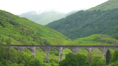 Historisches-Glenfinnan-Viadukt-Zwischen-Grüner-Landschaft-Und-Riesigen-Wäldern-Im-Hintergrund-An-Einem-Bewölkten-Tag