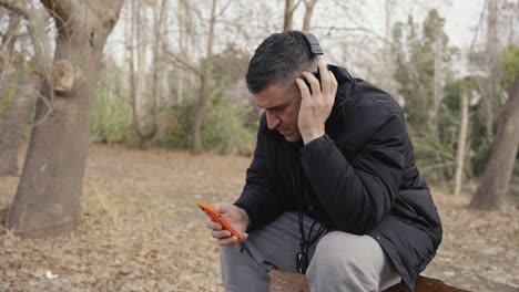 Ein-Mann-Sitzt-Im-Wald-Und-Scrollt-Durch-Sein-Handy,-Während-Er-Musik-über-Seine-Kopfhörer-Hört