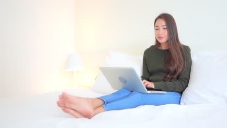 While-sitting-on-a-comfortable-bed,-a-young-woman-with-her-laptop-on-her-legs-inputs-data