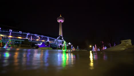 Niagara-Falls-Queen-Victoria-Place-Mit-Skylon-Tower-In-Der-Ferne-Nacht-Mit-Gefrorenem-Eis-Und-Schnee-Im-Winter-Festival-Der-Lichter-Weihnachtsbäume,-Die-Im-Winter-Auf-Eis-Reflektieren
