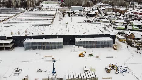 Modern-logistic-building-construction-site-with-Kaunas-city-behind-during-heavy-snowfall,-aerial-side-fly-view