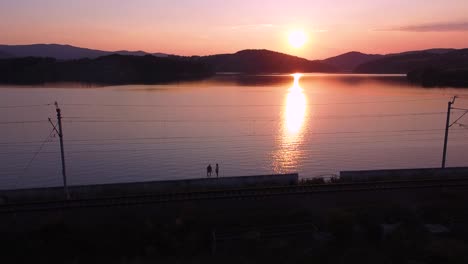 Aerial-shot-of-walking-couple,-railway-track,-clear-sky,-sunset-and-huge-orange-lake