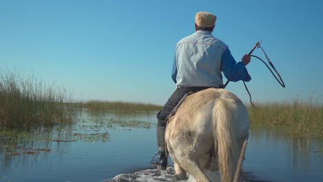 Rural-man-rides-white-horse-across-estuary-holding-riding-crop,-slow-motion