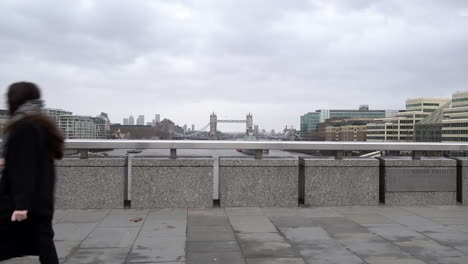 People-walk-across-London-Bridge-and-pass-Tower-Bridge-and-the-Thames-river-in-the-background-on-a-cold,-grey-and-windy-day-in-slow-motion