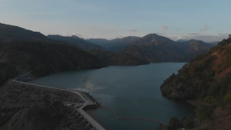 water-reserve-mountainous-region-Ambuklao-hydroelectric-Dam-revealing-lake-surrounded-by-trees-mountains-in-remote-area-Luzon,-Bokod,-Benguet,-Philippines-Wide-Aerial-reversing-away-revealing-road