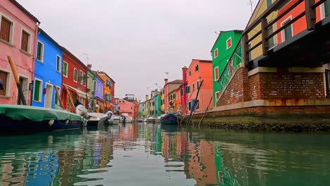 Water-surface-pov-view-of-Burano-picturesque-colored-houses-and-canal-with-moored-boats,-Italy
