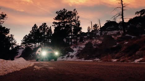 Stunning-tracking-shot-of-a-blue-SUV-with-bright-headlights-driving-along-a-snowy-dirt-mountain-road-during-sunset
