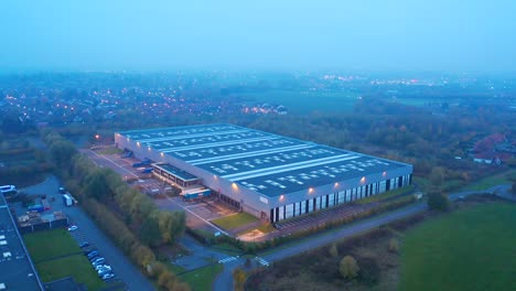 Warehouse-at-dusk:-Aerial-moving-back-up,-with-the-lights-of-the-nearby-city-and-roads