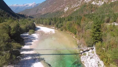 drone-tilt-up,-man-walking-across-rope-bridge-above-beautiful-crystal-clear-Alpine-river