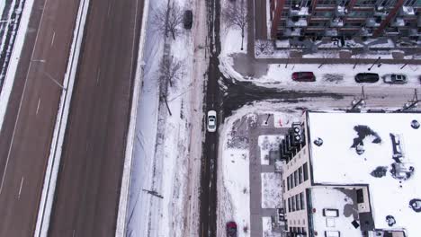 White-Car-Driving-Down-Snowy-Roads-in-a-Downtown-City-Past-a-Highway-and-Buildings