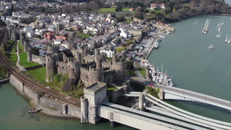 An-wider-aerial-view-of-Conwy-Castle-on-a-sunny-day,-flying-left-to-right-around-the-castle-while-zooming-out-with-the-town-in-the-background,-North-Wales,-UK