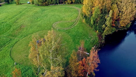 Aerial-top-down-shot-of-grass-field-with-barrel-sauna-next-to-natural-lake-during-daytime---Idyllic-landscape-in-nature