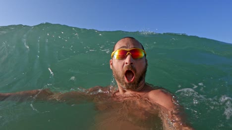 Active-bearded-man-with-yellow-sunglasses-having-fun-splashing-with-big-sea-waves-breaking-on-his-head