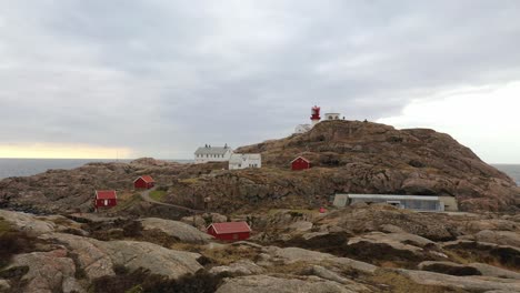 Lindesnes-lighthouse-forward-moving-low-altitude-aerial---Cliff-foreground-in-lower-frame-with-lighthouse-tower-and-museum-buildings-in-background-against-dramatic-sky---Norway
