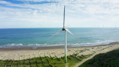 Aerial-view-of-rotating-wind-turbine-on-sandy-beach-and-wind-farm-in-background-standing-in-water-of-ocean
