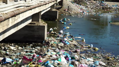 Local-river-filled-with-plastics-and-other-garbage-near-concrete-bridge,-static-view