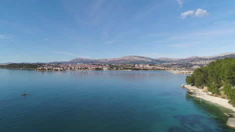 Tropical-coastline-and-majestic-city-of-Split-in-distance,-aerial-ascend-shot-over-seawater