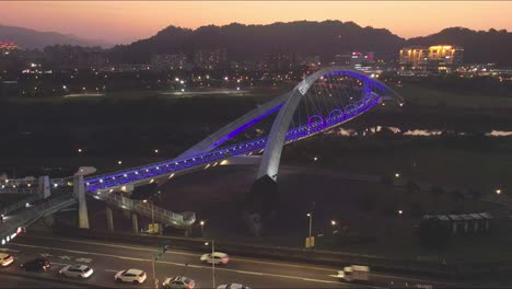 Vista-Aérea-Del-Tráfico-En-La-Autopista-Y-El-Puente-De-Iluminación-Azul-Con-Gente-Caminando-En-La-Ciudad-De-Taipei-Durante-La-Hora-Dorada