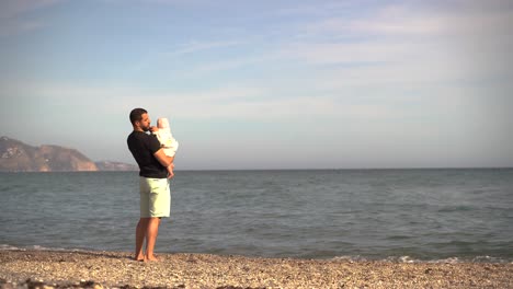 Vater-Hält-Liebevoll-Kleines-Baby,-Während-Er-Am-Strand-Auf-Das-Meer-Blickt