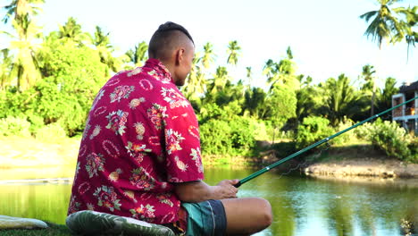 Young-adult-fishing-near-small-tropical-lake-with-palm-trees-in-background,-back-view
