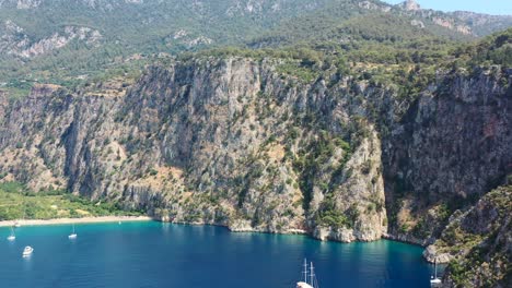 aerial-drone-panning-up-towards-the-large-green-mountains-of-Butterfly-Valley-while-boats-are-anchored-in-the-blue-ocean-of-Fethiye-Turkey