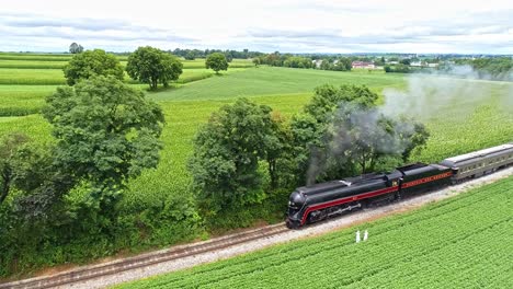 Eine-Luftaufnahme-Von-Amish-Ackerland-Mit-Einem-Einzigen-Eisenbahngleis-Und-Einem-Dampf-Personenzug,-Der-Sich-Nähert