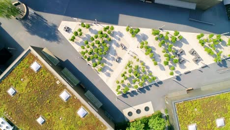Aerial-birdseye-view-of-a-modern-plaza-with-angular-shapes,-small-green-trees,-people,-and-surrounded-by-shops-with-greed-roofs-pull-back