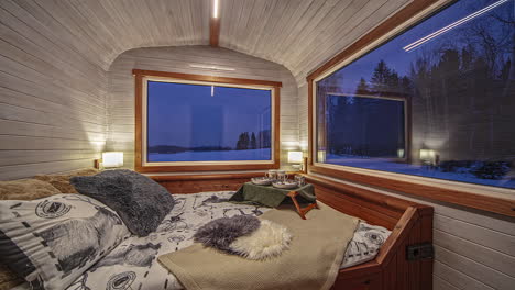 Static-view-of-a-cozy-bedroom-in-a-wooden-cabin-with-huge-glass-window-looking-outside-to-the-snow-covered-countryside-at-dawn-in-timelapse