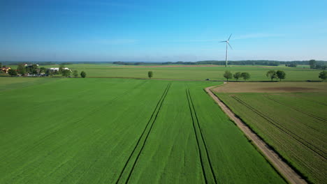 Aerial-view-crossing-rural-countryside-farmland-towards-spinning-wind-turbine-on-lush-agricultural-pasture
