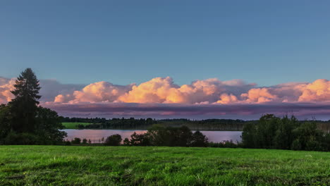 Timelapse-video-of-fast-moving-white-puffy-cloudscape-on-clear-blue-sky-over-rural-landscape-with-green-grasslands-and-lake-in-the-evening-time