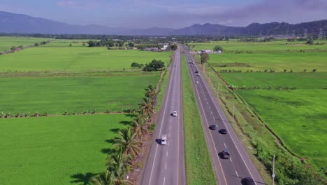 Aerial-flyover-motorway-with-traffic-on-Bonao-surrounded-by-green-growing-rice-fields-and-dark-clouds-in-backdrop