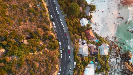 top-down-view-of-cars-driving-on-Clifton-Beach-Road-during-sunset-in-Cape-Town,-aerial
