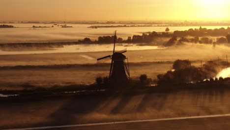 Low-aerial-shot-of-a-dramatic-silhouetted-windmill-and-farm-fields-with-a-light-fog-clinging-to-the-ground-in-early-morning-bright-orange-sun
