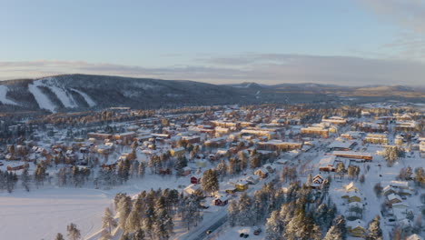 Aerial-view-across-vast-Scandinavian-snow-covered-settlement-surrounded-by-forested-mountain-landscape-under-sunrise-skyline