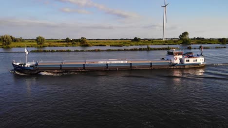 Aerial-Port-Side-View-Of-Amare-Inland-Motor-Freighter-Travelling-Along-Oude-Maas-Against-Still-Wind-Turbines-In-The-Background