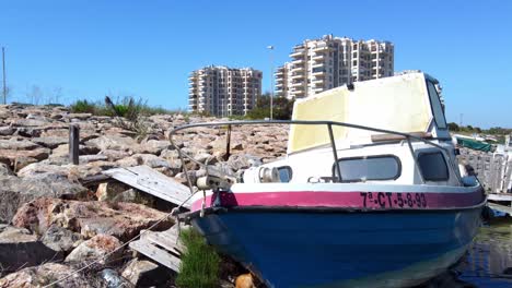 An-old-motorboat-tied-along-a-rocky-shoreline-then-aerial-ascent-to-view-a-apartments-and-condos-in-the-background