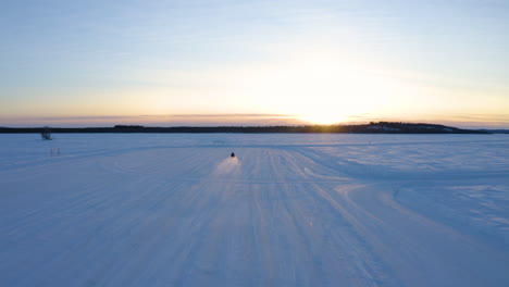 Aerial-view-lone-snowmobile-driver-travelling-fast-across-flat-snowy-Nordic-winter-wilderness-towards-sunrise