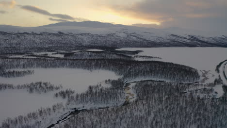 Aerial-view-across-snow-covered-Scandinavian-mountain-range-woodland-wilderness-under-sunrise-skyline