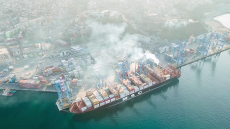 Aerial-hyperlapse-of-container-cargo-ship-docked-near-cranes-being-loaded-in-Valparaiso-Sea-Port,-vapor-in-air,-Chile