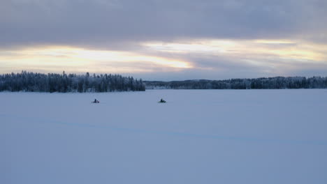 Aerial-view-pair-riding-snowmobiles-across-flat-snowy-Nordic-winter-wilderness-at-sunrise